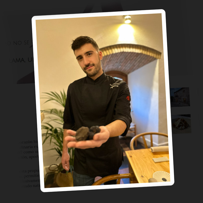 A portrait of Chef Mario Clemente in a dark, atmospheric kitchen, illuminated by a warm, orange glow from a nearby wood fire, creating deep shadows and highlighting his focused expression.
