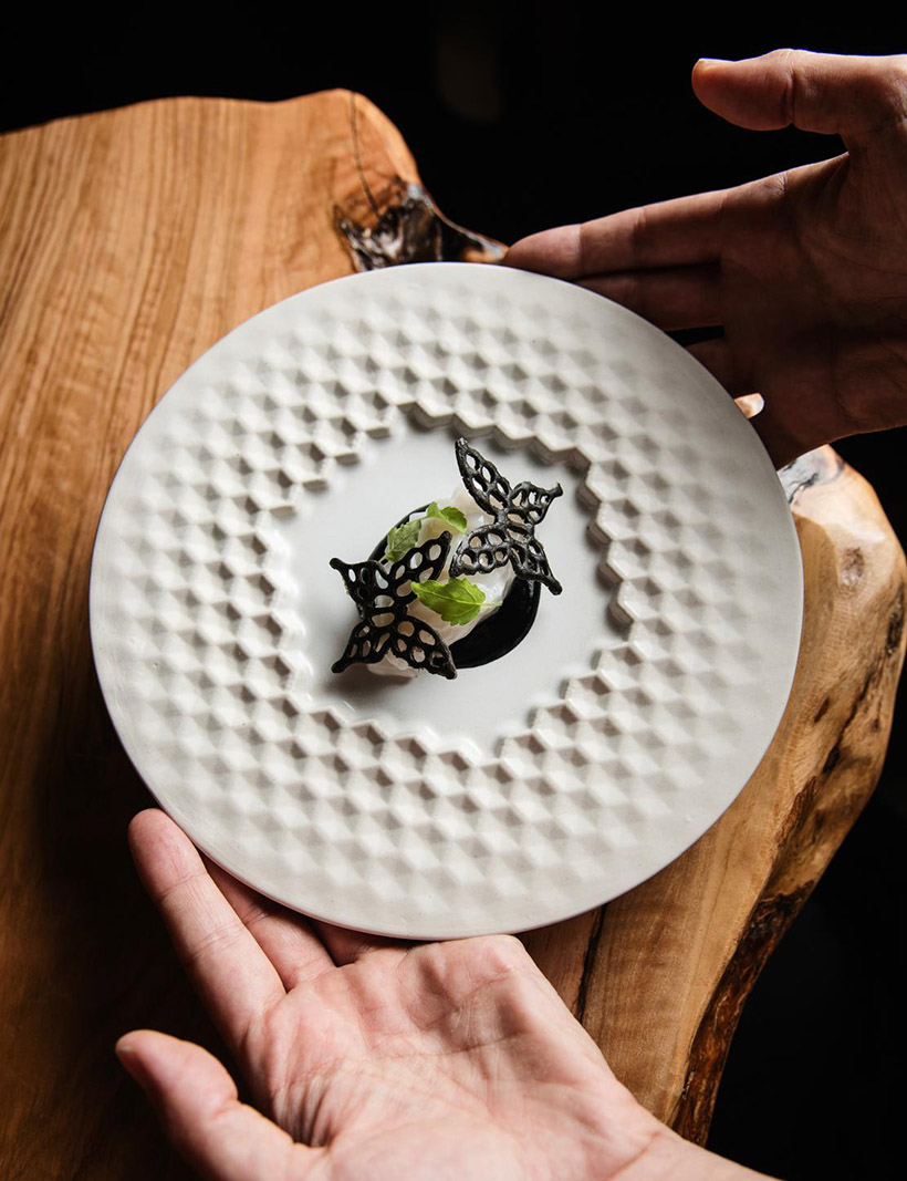 An exquisite, minimalist plated dish featuring wood-fired vegetables and smoked essence. The presentation is artistic and contemporary, set against a dark ceramic plate that blends into the charcoal background. Subtle rim lighting highlights the textures of the food, creating a luxurious and reverent culinary atmosphere. The color palette is dominated by deep tones with a single vibrant ember-colored accent.